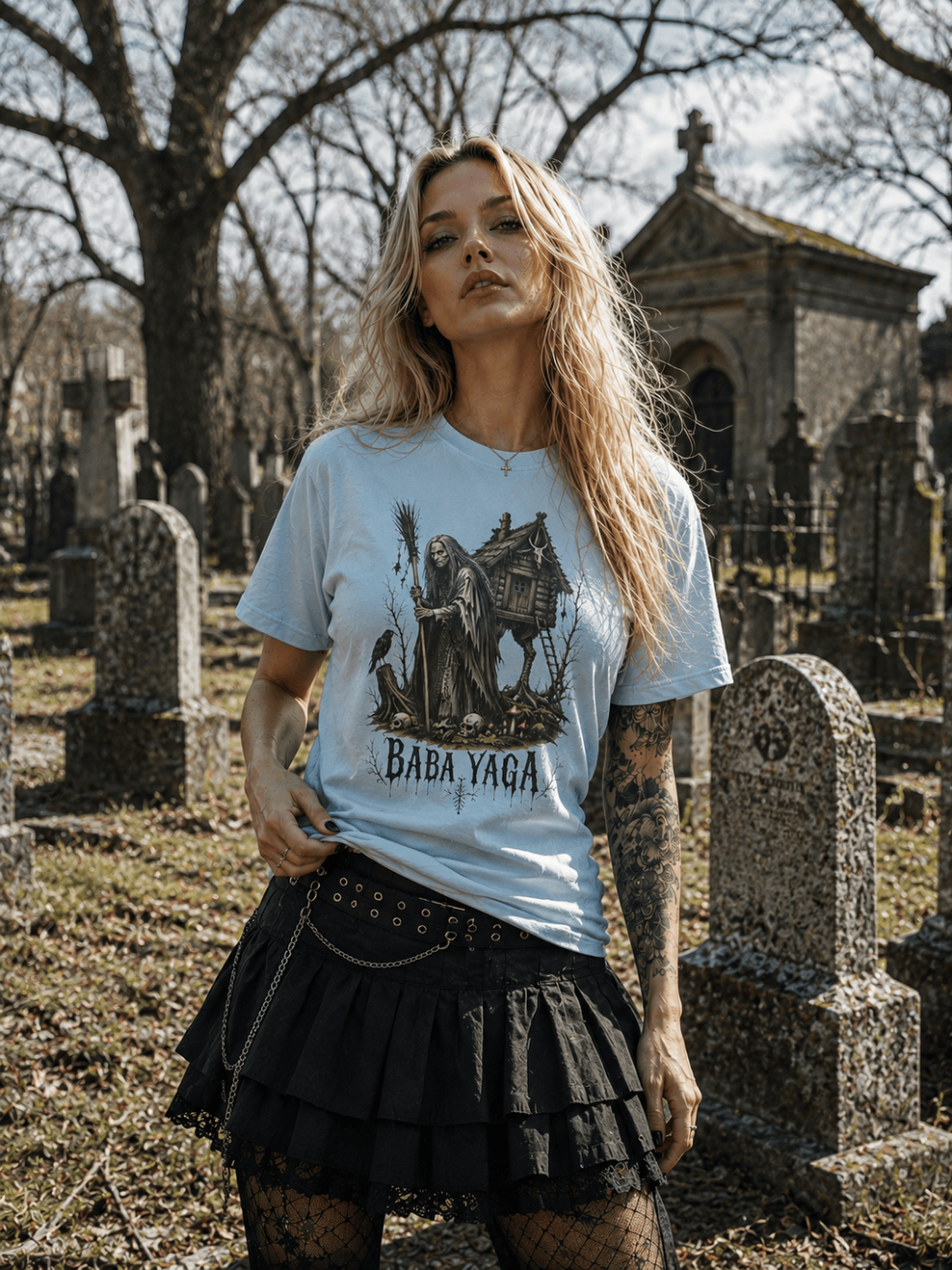 A woman standing in a cemetery wearing a light blue graphic t-shirt with 'Baba Yaga' printed on the front, paired with a black pleated skirt.