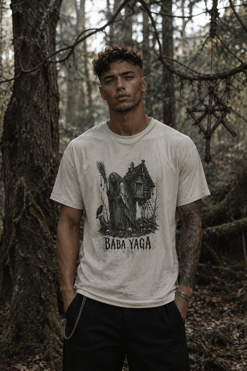 A man wearing a cream-colored graphic t-shirt with a black illustration of a haunted house and the text 'BABA YAGA', standing in a dense, wooded forest.