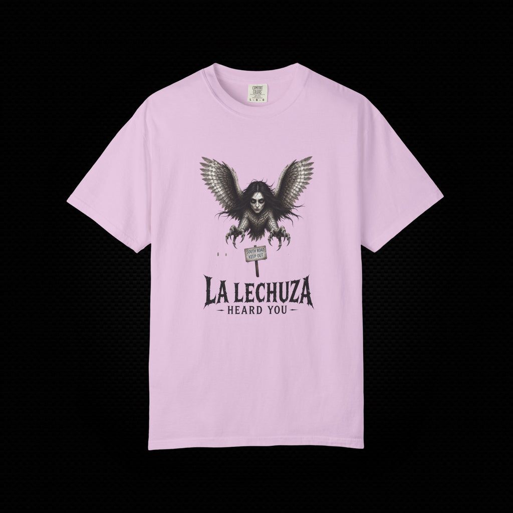 A light pink short-sleeve t-shirt with a black graphic of a winged witch holding a sign that reads 'LA LECHUZA - HEARD YOU -' on the front, displayed against a solid black background.
